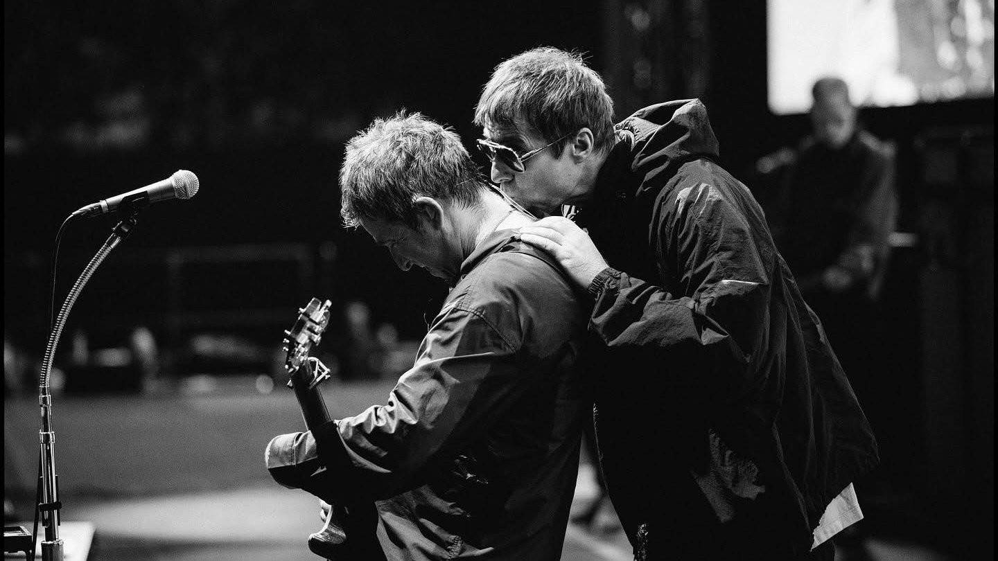 Liam Gallagher sahnede gitar çalan Noel Gallagher'ın omzuna sarılıyor